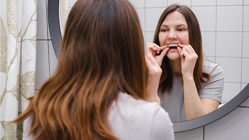 A woman in front of a mirror in the bathroom holding invisible plastic teeth aligners in hands and putting on braces. Beautiful and healthy smile.