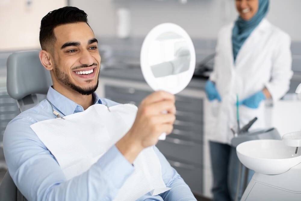 Happy Handsome Arab Man Looking To Mirror After Teeth Treatment In Modern Clinic, Middle Eastern Male Patient Sitting In Chair In Stomatological Cabinet And Enjoying His New Smile, Closeup
