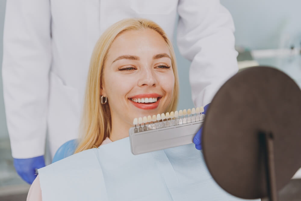 Close up young smiling happy caucasian woman choose veneers enamel color palette look at mirror sit at dentist office chair indoor cabinet near stomatologist Healthcare oral enamel aesthetic treatment
