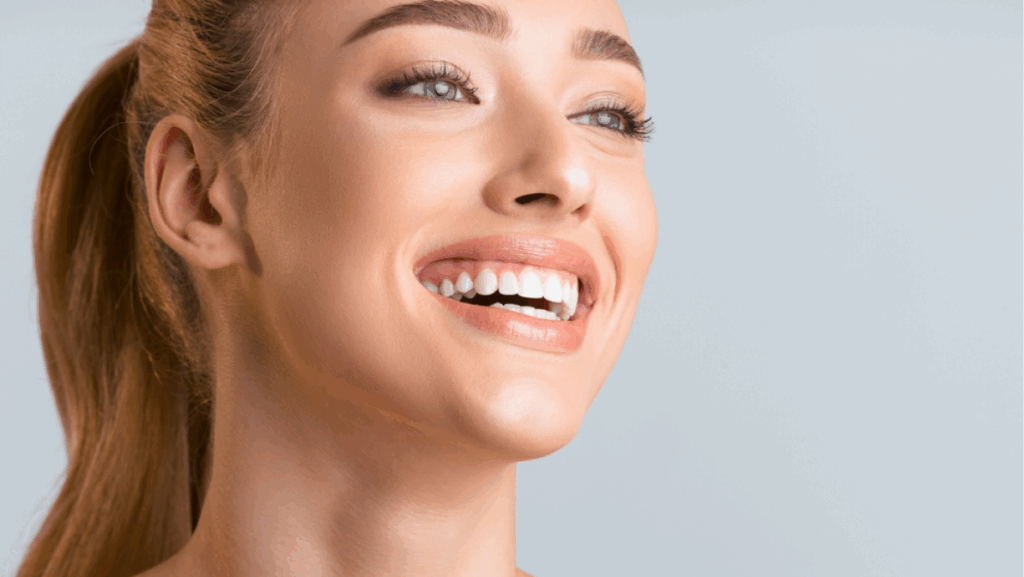A close-up portrait of a young woman with light brown hair pulled back in a ponytail, smiling brightly. Her skin is fair and glowing, and her teeth are white and bright, copy space
