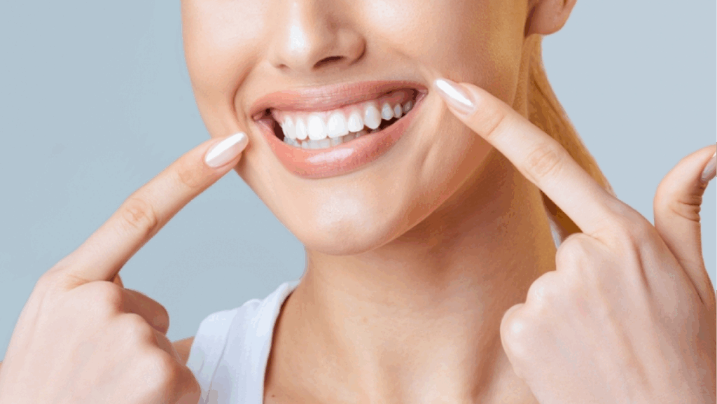 This image is a close up of a woman face. Her fingers are resting on her cheeks, and she is smiling widely, showing off her bright white teeth, cropped