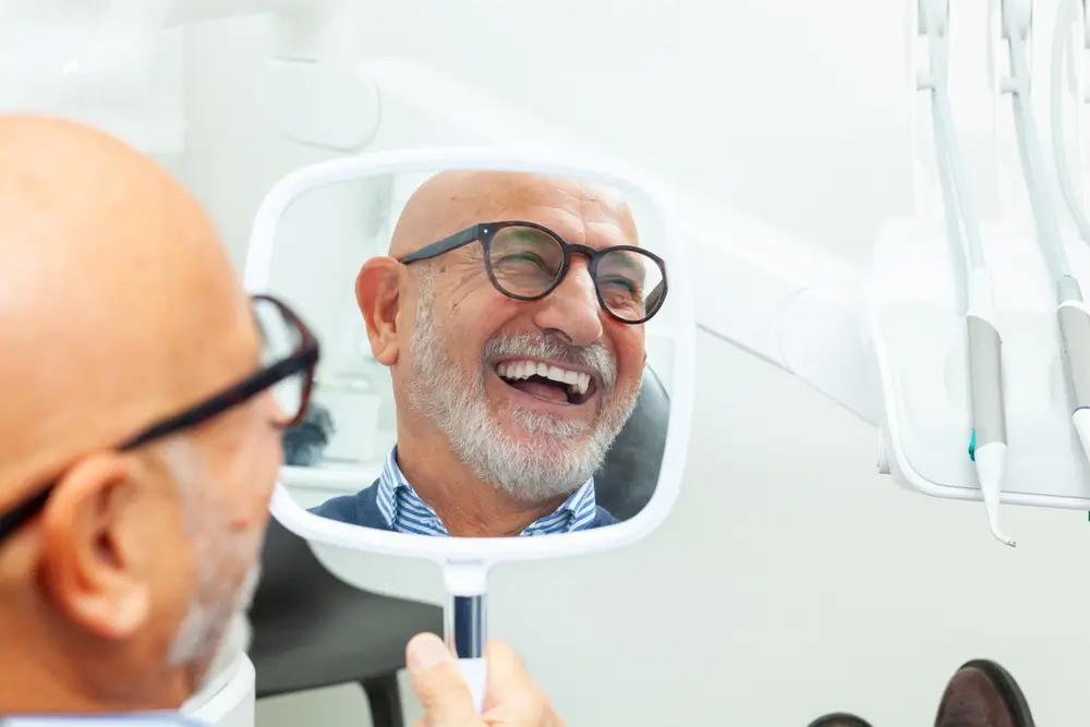 Senior patient admiring his perfect smile after a dental implant treatment with a mirror in his hand