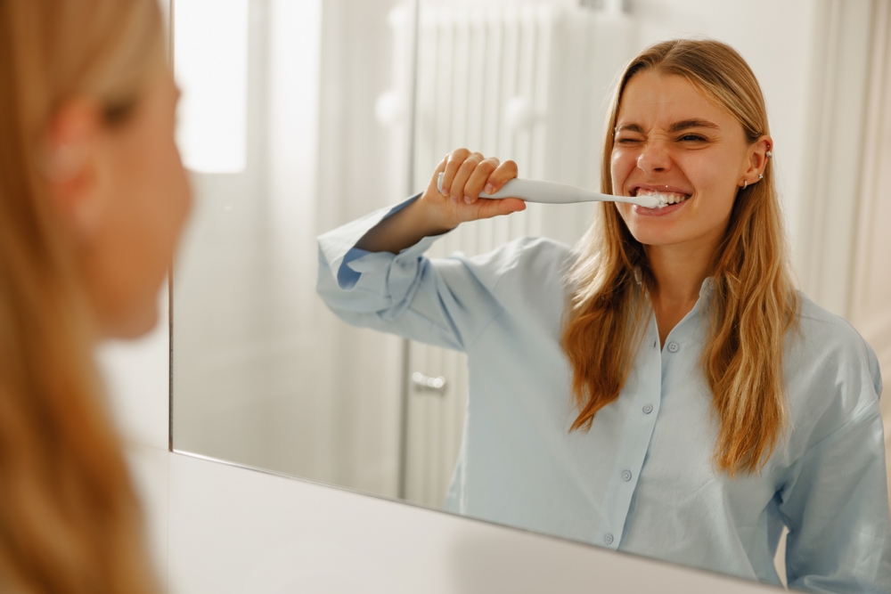 A young woman happily enjoys her refreshing morning routine, brushing her teeth and smiling brightly in the mirror